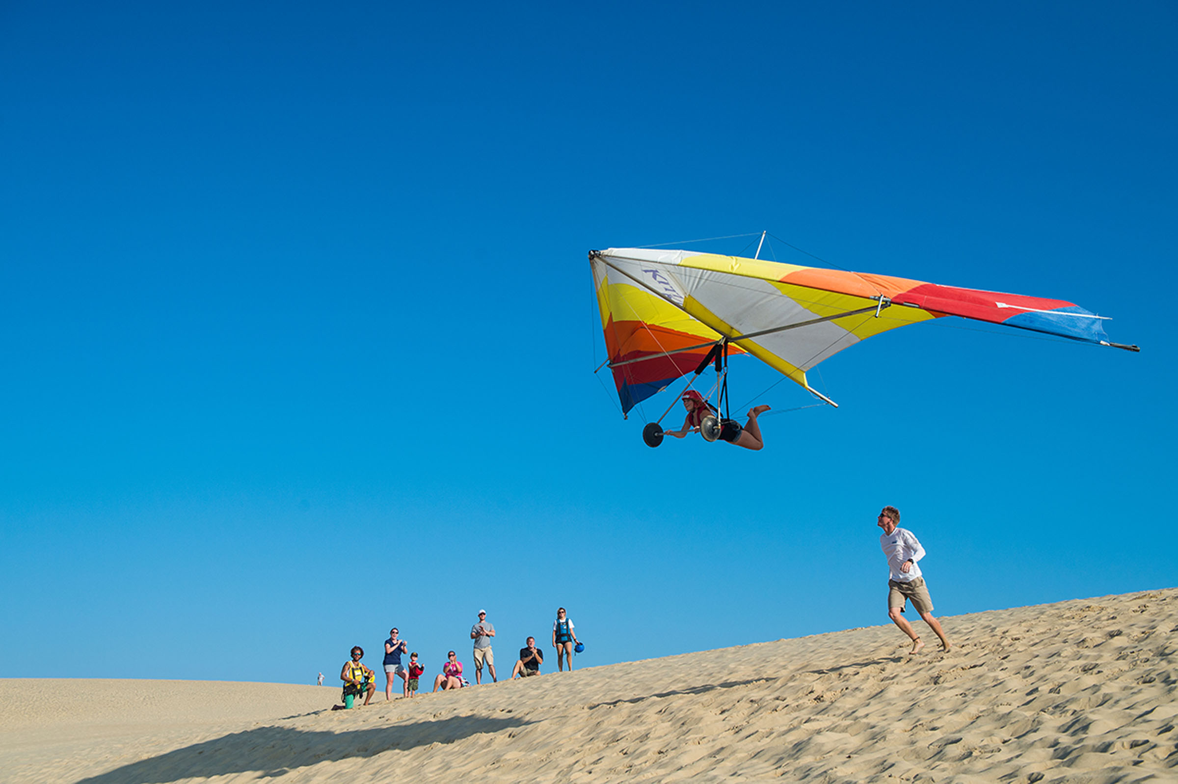 A hang glider flies low over sandy dunes with a small group of people watching below.