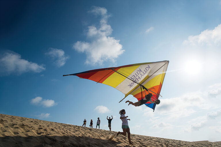 A hang glider flies low over a sandy dune with several people watching nearby.