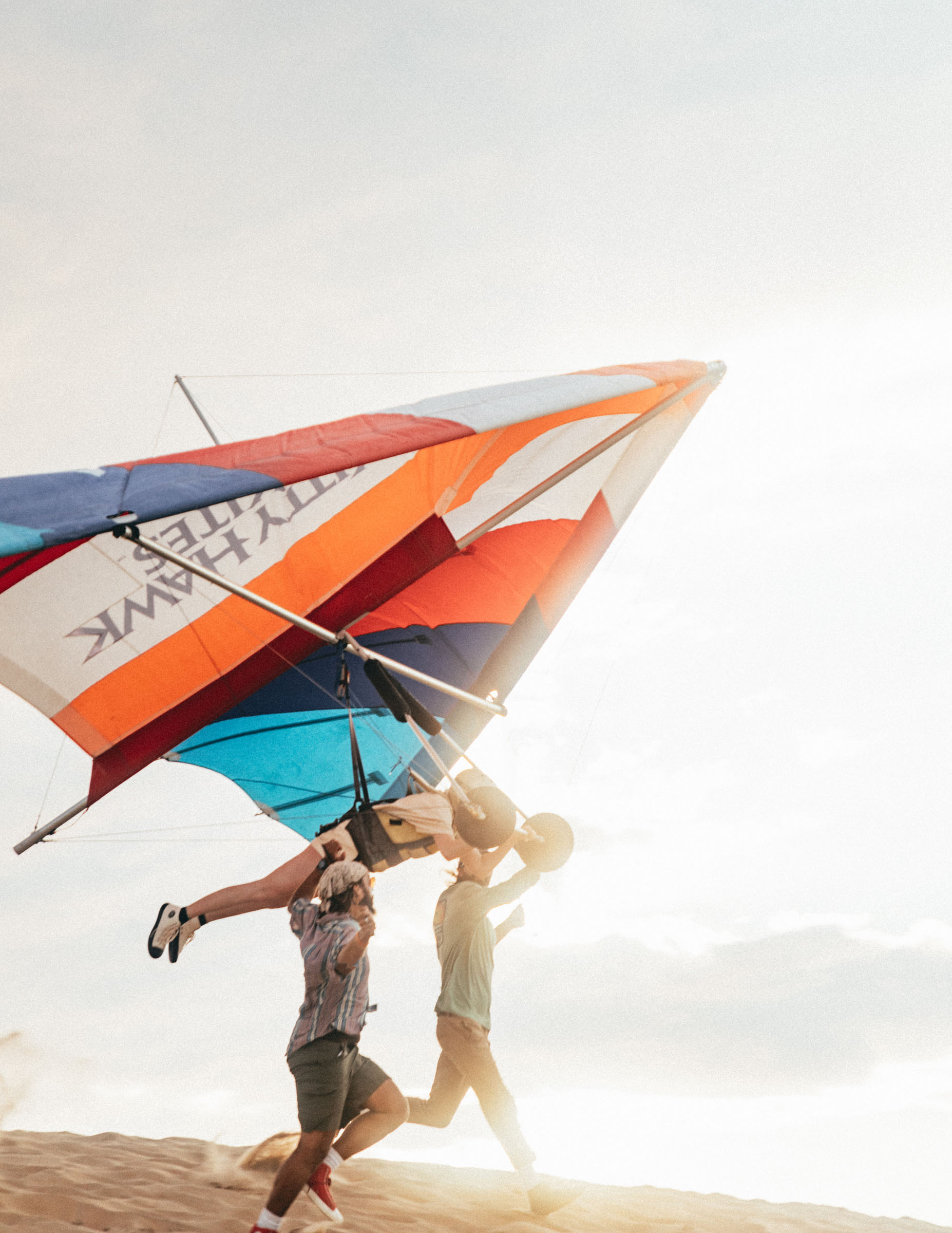 Two people run across a sand dune while carrying a colorful hang glider.