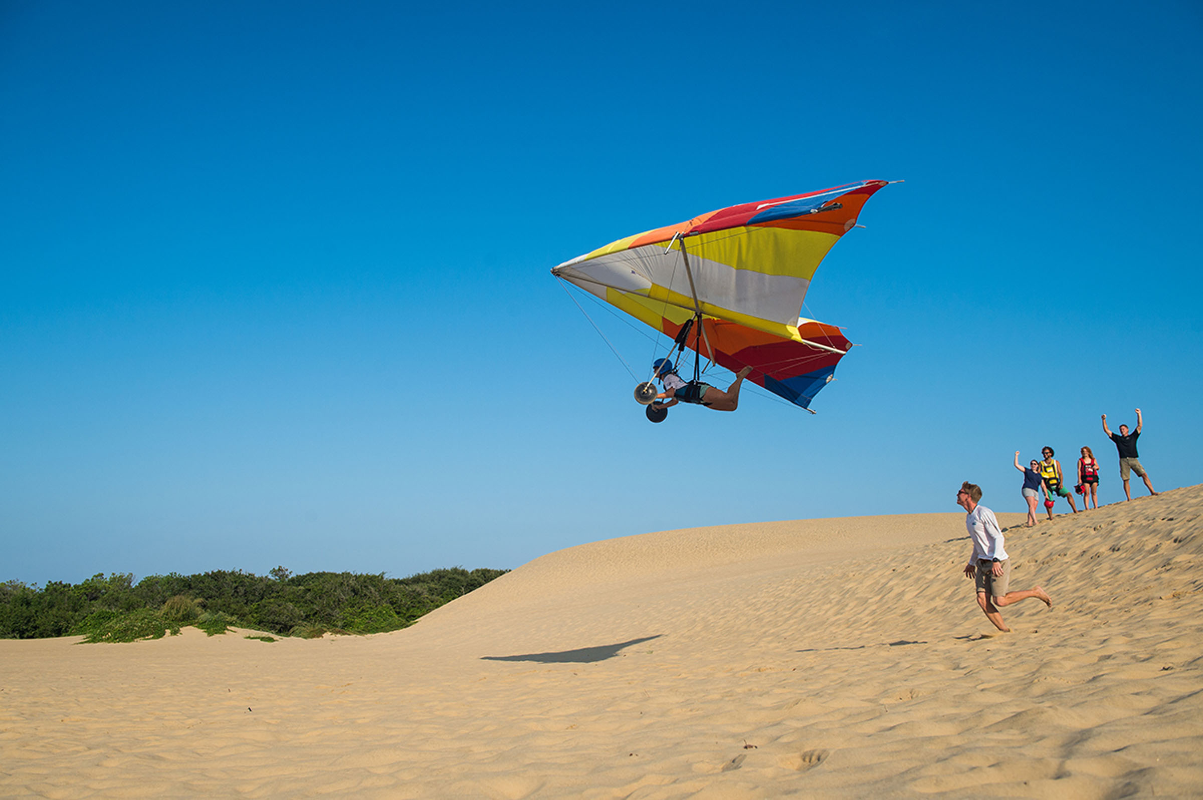 A hang glider flies above sand dunes while several people watch nearby.