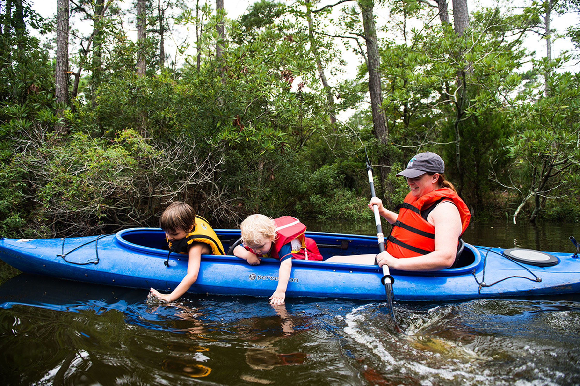 A woman and two children ride in a blue kayak on a calm wooded waterway.
