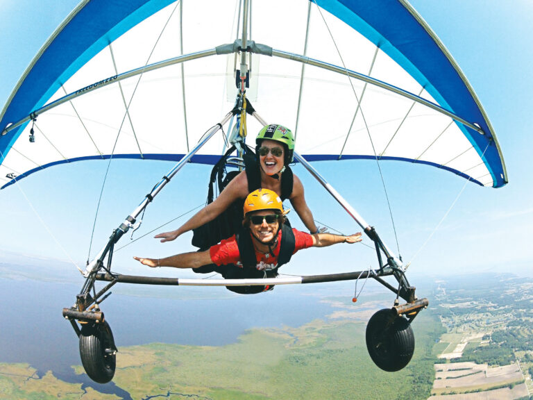 Two people smile while tandem hang gliding high above the coastline and marshland.