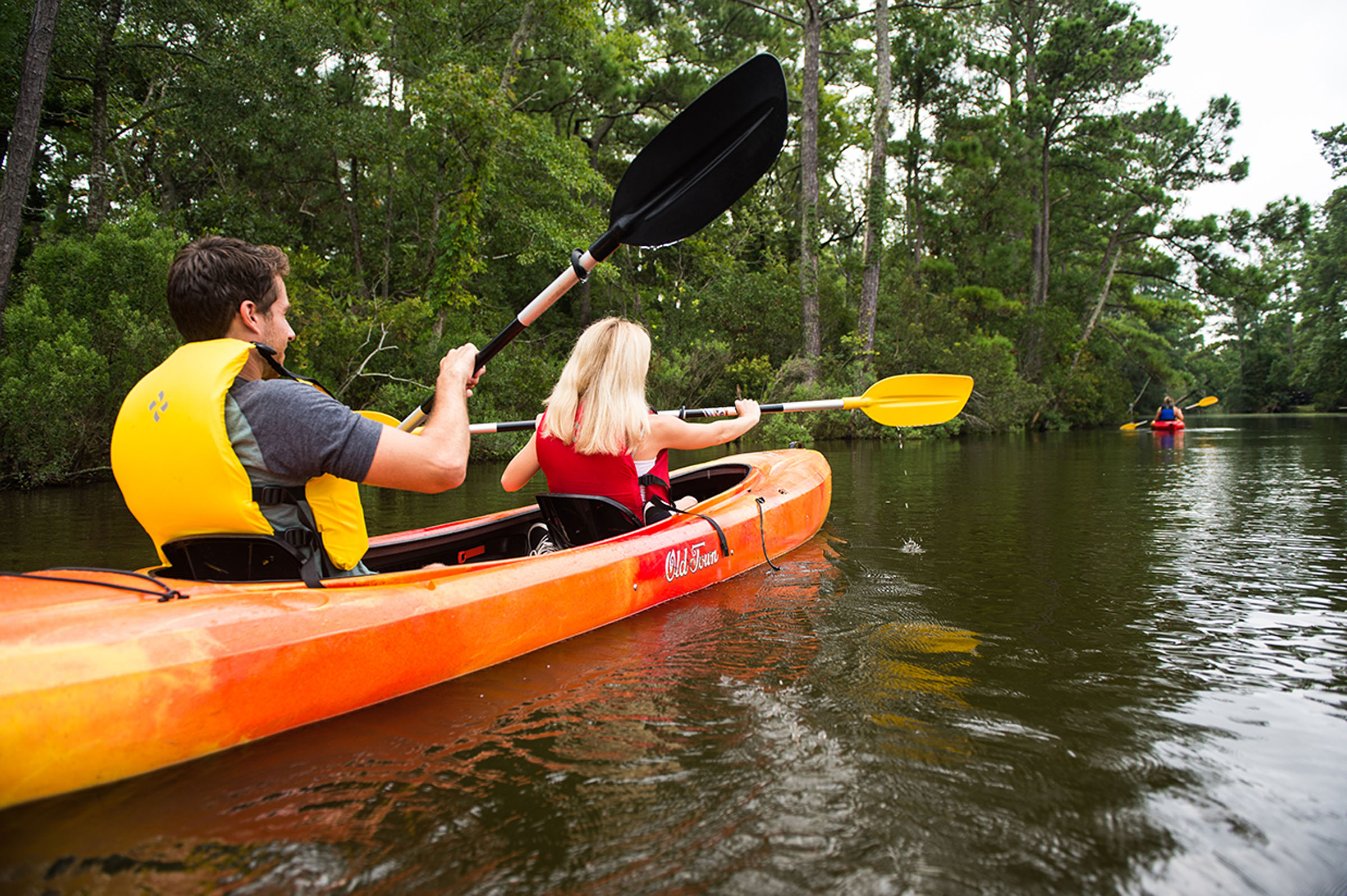 Two people paddle an orange kayak along a calm wooded waterway.