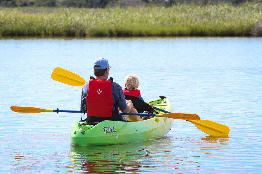 Two people paddle a green kayak on calm water with marsh grass in the background.