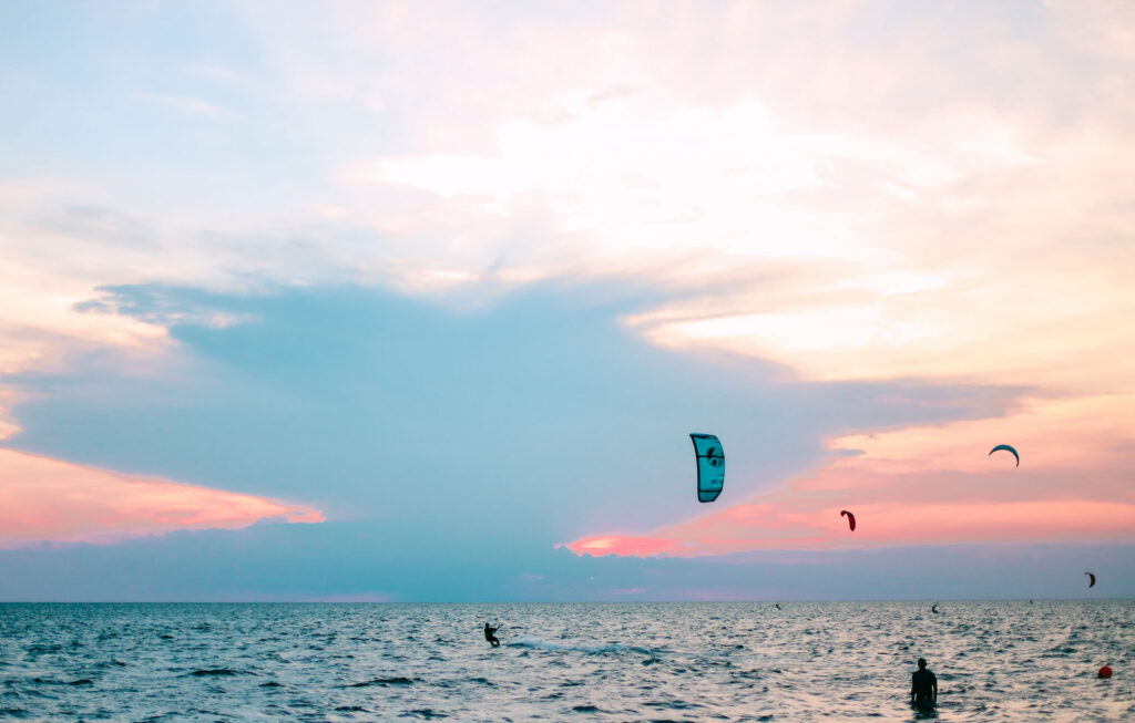 Kiteboarders ride across the ocean under a pastel sunset sky.