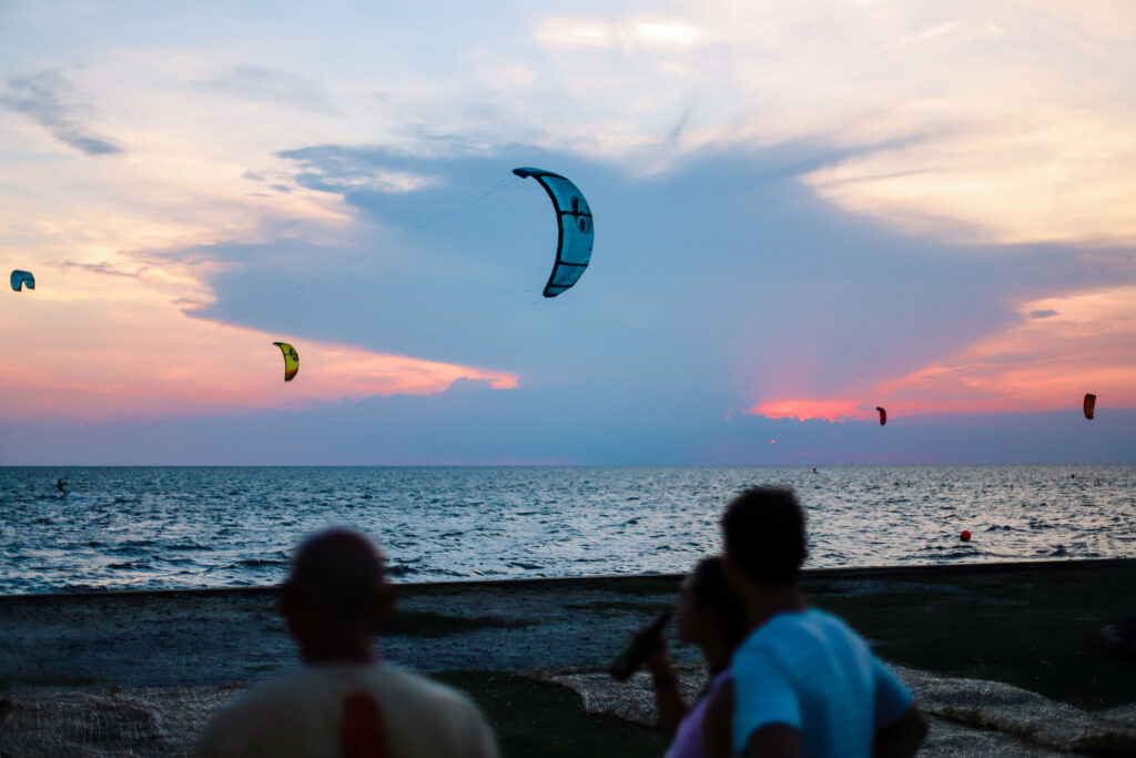 Kiteboarding kites fly over the ocean at sunset with people watching from the beach.