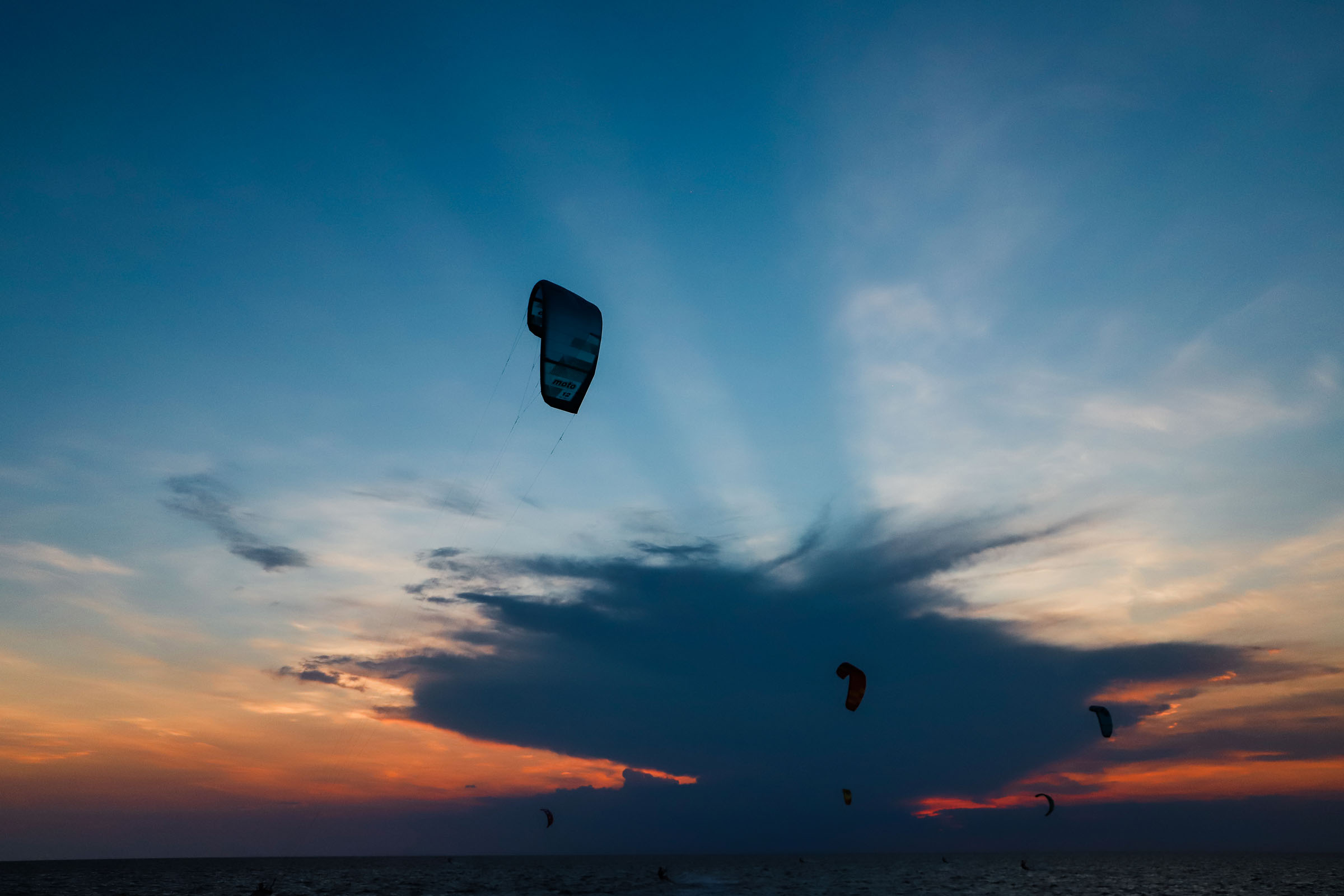 Kiteboarding kites are silhouetted against a colorful sunset sky over the water.