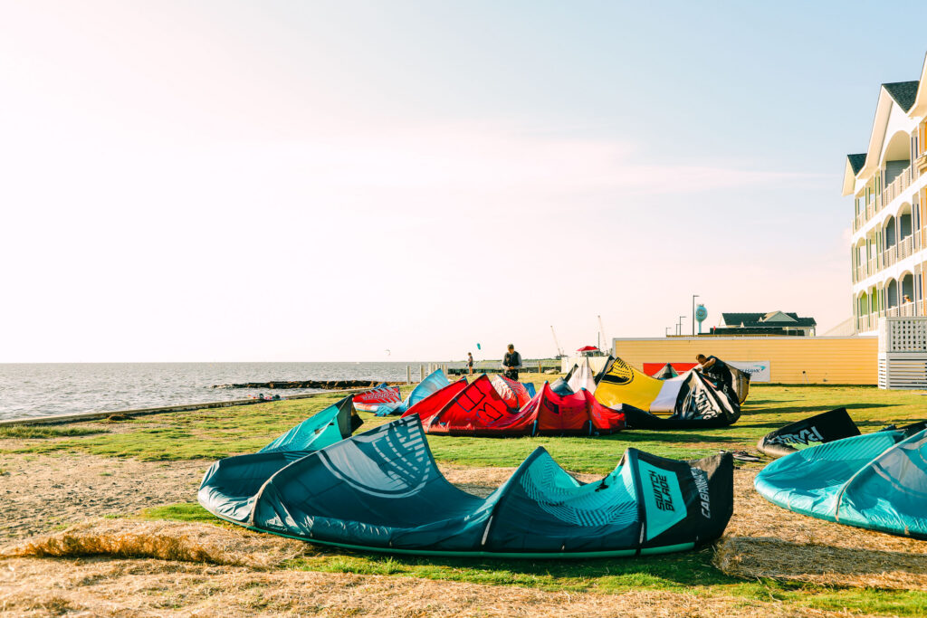 Colorful kiteboarding kites are laid out on a grassy waterfront near the beach.