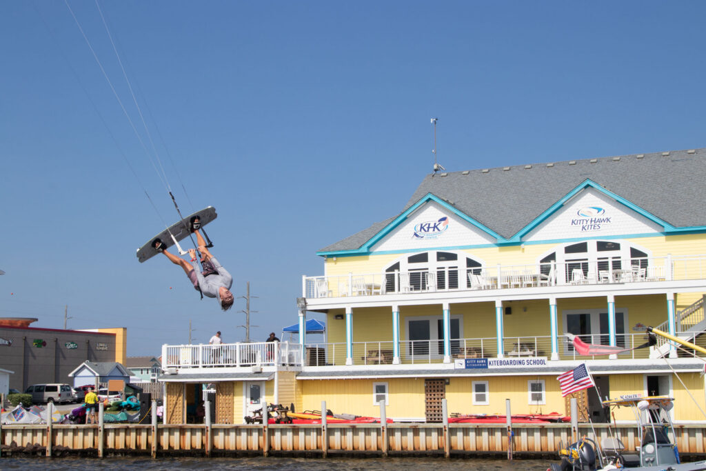 A kiteboarder flips in the air near a waterfront kiteboarding school.
