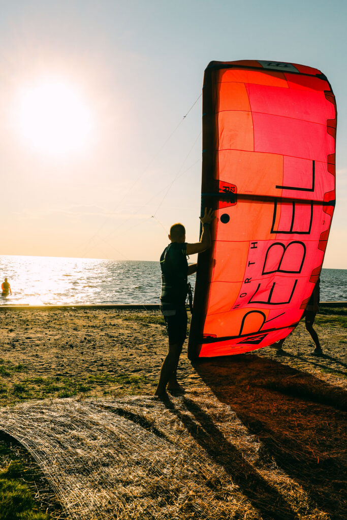 A person stands on a beach holding a large red kite beside the water at sunset.